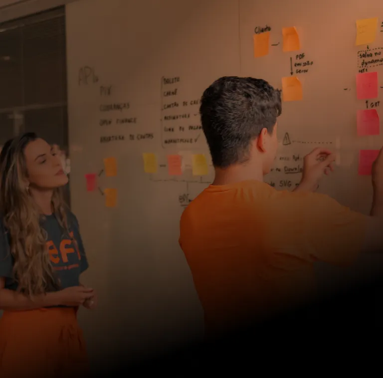 Colaborador do Efí Bank, de costas, vestindo uma camisa laranja e colando adesivos em um quadro de planejamento